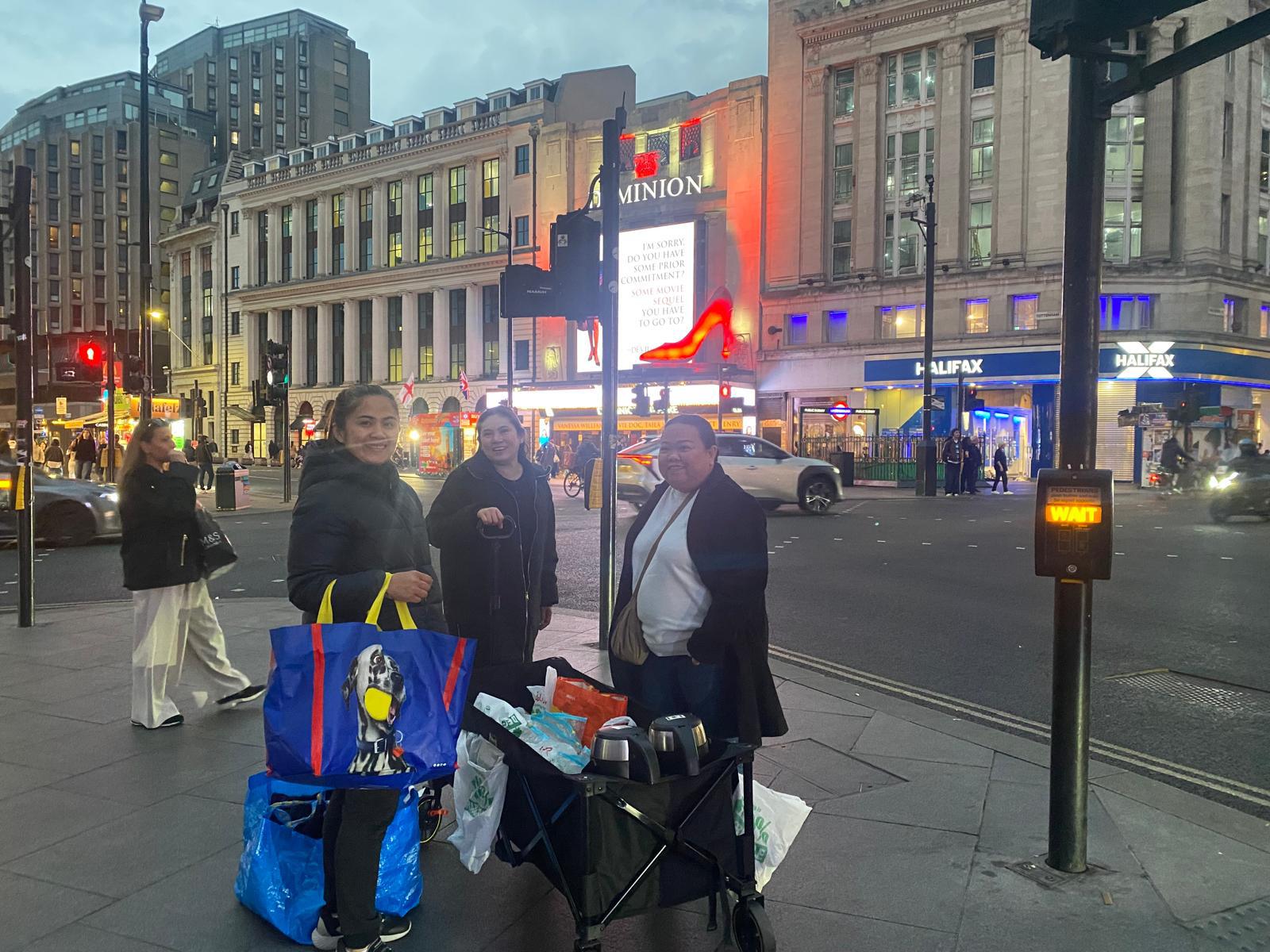 Three FWA-UK Outreach Committee volunteers pause with their supply wagon near Tottenham Court Road, with the Dominion Theatre visible behind them.