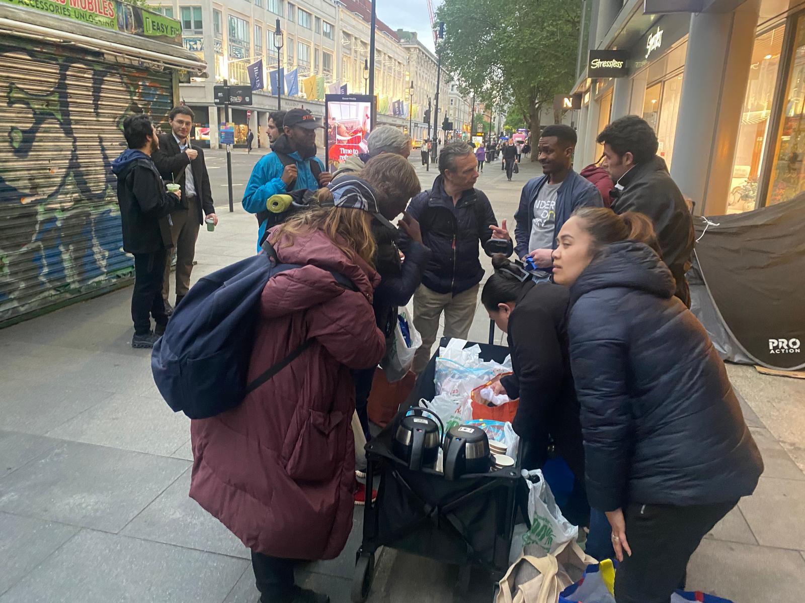 Volunteers and community members gathering around a supply cart on Oxford Street, receiving hot drinks and food parcels during the FWA-UK and Under One Sky outreach session.